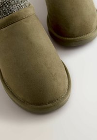 Grønne ruskindsko med rundede tæer, der har en struktureret strikket kant. Glat overflade og kontrasterende syning langs sålen.