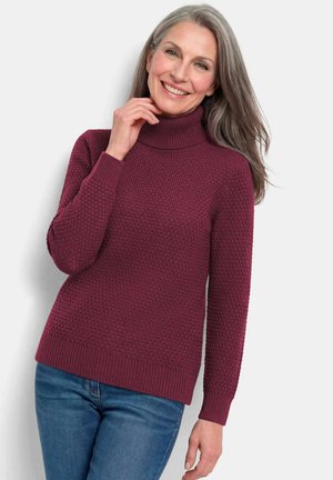 Frau mit langen grauen Haaren, lächelnd, trägt einen strukturierten dunkelroten Rollkragenpullover und blaue Jeans, steht vor einem einfachen hellen Hintergrund.