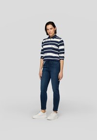 Navy- und weiß gestreiftes Polohemd mit Kragen, kombiniert mit dunkelblauen eng anliegendem Jeans und weißen Sneakers, steht vor einem neutralen Hintergrund.