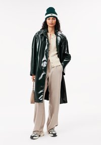 Cappotto in vinile verde lucido, maglione a trecce crema, pantaloni beige a gamba larga, sneakers grigie, berretto lavorato a maglia verde e bianco, piccola borsa beige.