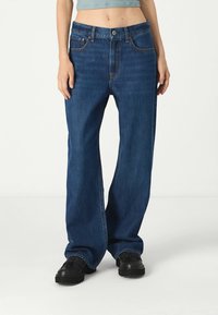 Pantalones vaqueros de denim azul de pierna ancha con una cintura alta, que cuentan con bolsillos delanteros, cierre de botón y costuras en contraste. Llevados con zapatos negros.