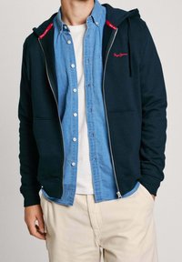 Sudadera con cremallera navy y cordones, con detalles rojos en el cuello y un logo. Llevada sobre una camisa de botones azul claro.