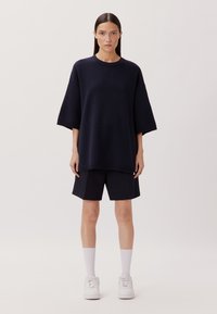 Maglione strutturato blu navy oversize con maniche ampie, abbinato a pantaloni scuri e calze bianche, indossato con sneakers bianche. Design semplice e rilassato.