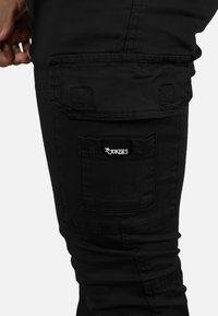 Pantalones cargo negros de algodón, con un gran bolsillo lateral y un pequeño parche con la marca "The Jokers". Corte ajustado con puños elásticos.