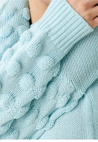 Pull tricoté bleu clair avec motifs en relief texturés sur un côté et poignets côtelés. Fil doux pour une sensation de confort.