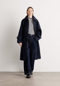 Lang, mørk, oversized frakk laget av teksturert stoff, med bred krage, knappelukking og frontlommer, kombinert med stripete topp og vide bukser.