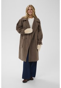 Smiling woman wearing oversized brown coat, white sweater, wide-leg jeans, brown boots, and beige fuzzy mittens standing on a plain gray background.