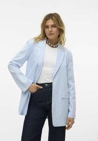 Blazer bleu clair avec une coupe décontractée, une poche unique et des manches longues. Porté sur une chemise blanche et assorti à un pantalon en denim foncé.