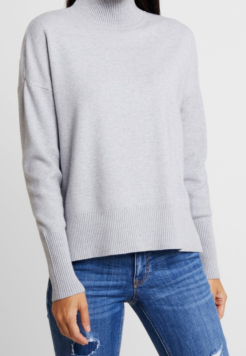 Femme portant un pull à col roulé gris clair avec des poignets et un ourlet côtelés, associé à un jean bleu déchiré.