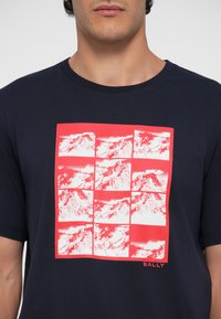 Tamnoplava majica s kratkim rukavima koja sadrži crvenu grafiku planinskog krajolika raspoređenu u mrežu; okrugli izrez.