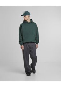 Grønn hettegenser med frontlomme, kombinert med løstsittende grå jeans og svarte sko. Capsen har en logo på forsiden.