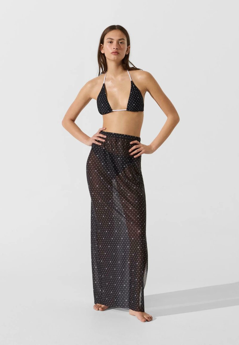 Femme debout pieds nus portant un haut de bikini noir à pois et une longue jupe transparente assortie, les mains sur les hanches, sur un fond uni.