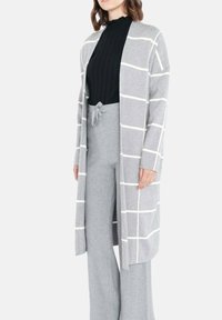 Long cardigan gris avec un motif à carreaux blancs, fait de tissu doux. Porté sur un haut noir côtelé et un pantalon large gris.