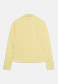 Pitkähihainen kauluspaita vaaleankeltaisena, valmistettu sileästä kankaasta. Siinä on raglan-hihat ja suora helma, jossa on hienovaraisia ompeluyksityiskohtia.