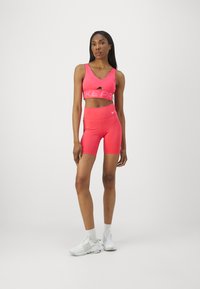 Roza športna modrček z izrezom, ki ima napis "NIKE PRO"; ujemajoče se roza hlače z visokim pasom; bele športne čevlje; gladka tekstura.