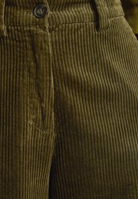 Olivengroene corduroy stof met verticale ribbels, knoopdetail op de taille en subtiele textuur overal.