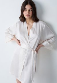 Accappatoio setoso di un leggero beige con scollatura a V, maniche ampie e dettagli in pizzo ai polsini. Presenta una cintura coordinata legata in vita.
