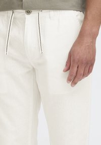 Gros plan sur la main d'une personne reposant à côté d'un pantalon blanc à cordon avec un t-shirt gris rentré à la taille.