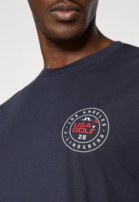 T-shirt azul-marinho apresenta um logótipo redondo com "USA GOLF" em vermelho e branco, com "LOS ANGELES" e "J. LINDEBERG" dispostos em padrão circular.