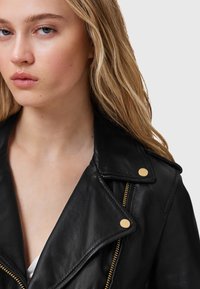 Giubbotto in pelle nera con colletto a punte, accenti in metallo dorato e chiusura a zip diagonale, che mostra una texture liscia e un design moderno.