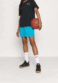 Person klädd i svart Nike-t-shirt, turkosa shorts, vita Nike-strumpor och svarta sneakers, som håller i en Nike-basketboll mot en vit bakgrund.