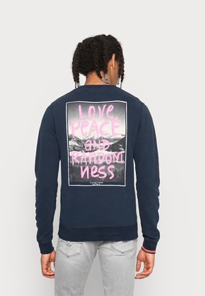 Hombre con cabello trenzado que lleva una sudadera azul marino con texto rosa "Amor, Paz y Aleatoriedad" sobre un fondo en blanco y negro de montañas.