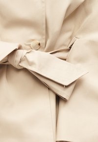 Υφασμάτινο παλτό τρεντσκόουτ σε μπεζ με δεμένη ζώνη και ένα ορατό μπεζ κουμπί κοντά στον κόμπο.