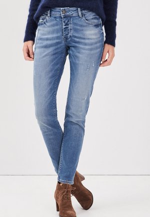 Jean skinny bleu avec des détails effilochés, présentant un design classique à cinq poches et une fermeture à bouton. Portés avec des bottines en suède marron.