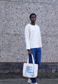 Lyse beige langærmet skjorte, denim jeans og en creme tote bag med blå tekst, sat op mod en struktureret grå væg.