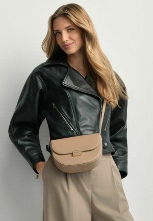 Kobieta w czarnej skórzanej kurtce i beżowych spodniach z beżową torebką na ramię, stojąca na tle jednolitego, jasnego tła.