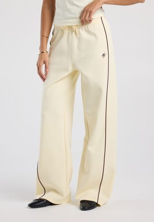 Vrouw draagt crèmekleurige wijde broek met zijbies en zwarte platte schoenen tegen een effen grijze achtergrond.