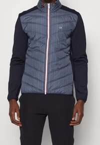 Quilted navy fabric jacket with a high collar, long sleeves, and contrast black arms. Features a front zipper and subtle logo detail.