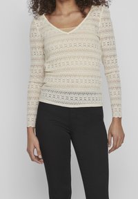 Lace långärmad topp i krämfärg, med V-ringning, intrikata blommönster och vågiga kanter, kombinerad med smala svarta byxor.