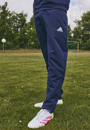 Person wearing navy blue Adidas track pants and white soccer cleats with pink and neon green accents standing on grass field.