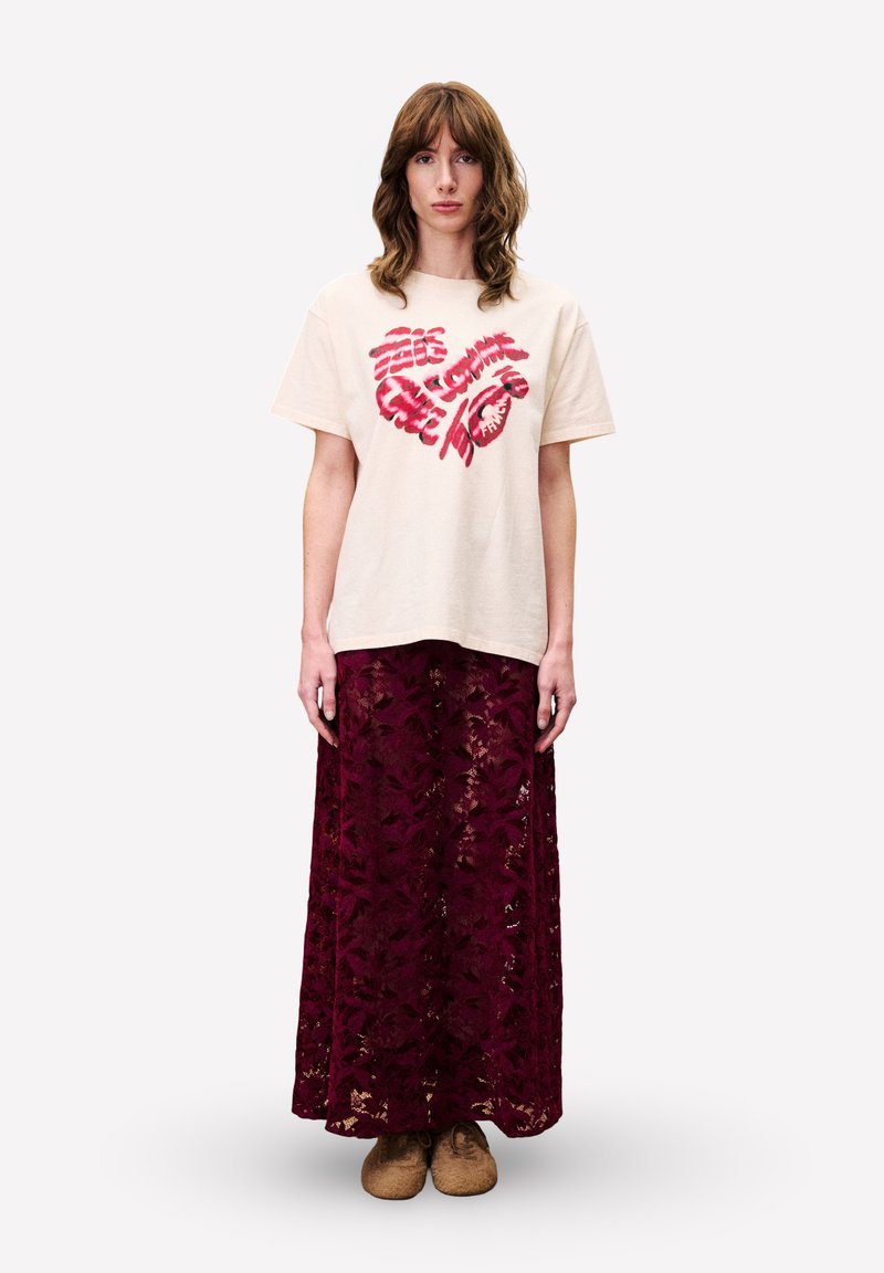 T-shirt beige en coton avec un motif graphique rouge, associé à une longue jupe en dentelle bordeaux. La jupe présente des motifs floraux et une coupe ample.