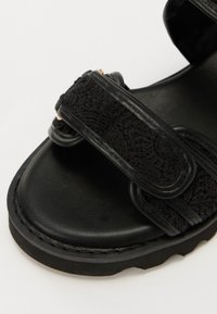 Sorte sandaler med teksturert stoffoverdel og glatte lærremmer. Har to justerbare remmer, kraftig såle og kontrasterende sømmer.