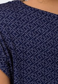 Marinblått tyg med små vita geometriska kvadratmönster som täcker axeln och överarmen på en person med ljusbrun hud.