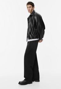 Jongeman in een zwarte leren jas, losse zwarte broek en zwarte laarzen, staande met de handen achter de rug tegen een witte achtergrond.