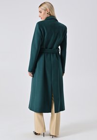Lange teal wollen jas met een ceintuur in de taille, reverskraag en een split aan de achterkant. Gekocht over beige broeken en zwarte puntige hakken.