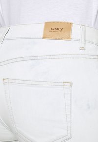 Jean de mezclilla azul claro con una etiqueta de cuero que dice "ONLY". La tela tiene una textura ligeramente desgastada con costuras visibles y un bolsillo trasero.