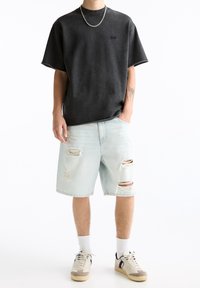 Camiseta oversized de color gris oscuro, de manga corta, con cuello acanalado; pantalones cortos de mezclilla desgastados de color azul claro; calcetines blancos; zapatillas crema y azul marino con cordones.