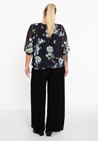 Sort blomsterbluse med gennemsigtigt stof og løs pasform, prydet med blå og grønne blomsterprint. Sammen med brede sorte bukser og hæle.