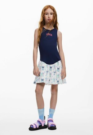 Canotta blu navy con etichetta "STITCH", gonna bianca svasata con motivo di granchi colorati, abbinata a sandali rosa e calzini azzurri.