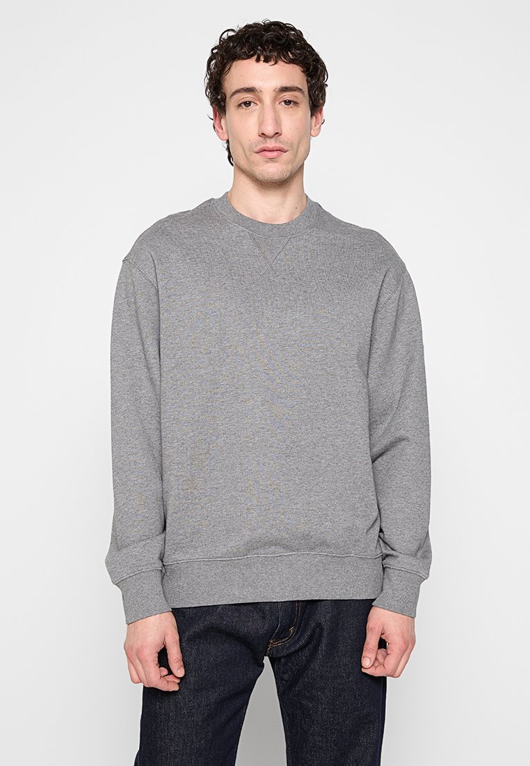 Hombre con cabello rizado corto, vestido con un suéter gris de cuello redondo y jeans oscuros, de pie frente a un fondo blanco.