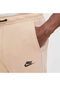 I pantaloni sportivi Nike beige presentano un tessuto morbido e leggero, tasche laterali, un logo nero e una sottile striscia grigia lungo la cucitura.