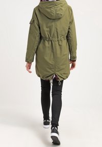 Oliwkowa parka z kapturem, ze sznurkiem w talii i kieszeniami na zamek, z delikatnie zaokrąglonym dołem i elastycznymi mankietami. Włożona z czarnymi legginsami.
