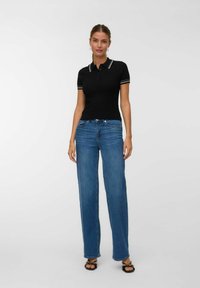 Kvinna står i blå raka jeans, svart kortärmad stickad polo med vit kant och svarta sandaler med remmar mot en enfärgad bakgrund.