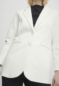 Bílý blazer z texturované látky, s klopovým límcem, dvěma knoflíky a bočními kapsami s upraveným střihem.