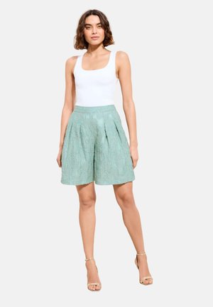 Kvinde iført hvid ærmeløs top, mintgrønne, højtaljede, texturerede shorts og beige ankelstroppede hæle, stående med hænderne langs siden.
