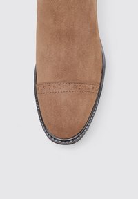 Zapato de ante marrón con punta redondeada, que presenta una banda cosida en la parte frontal y una suela negra texturizada con un borde decorativo.